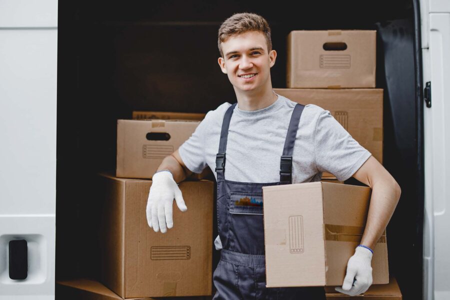 A young handsome smiling worker wearing uniform is standing next to the van full of boxes holding a box in his hands. House move, mover service.