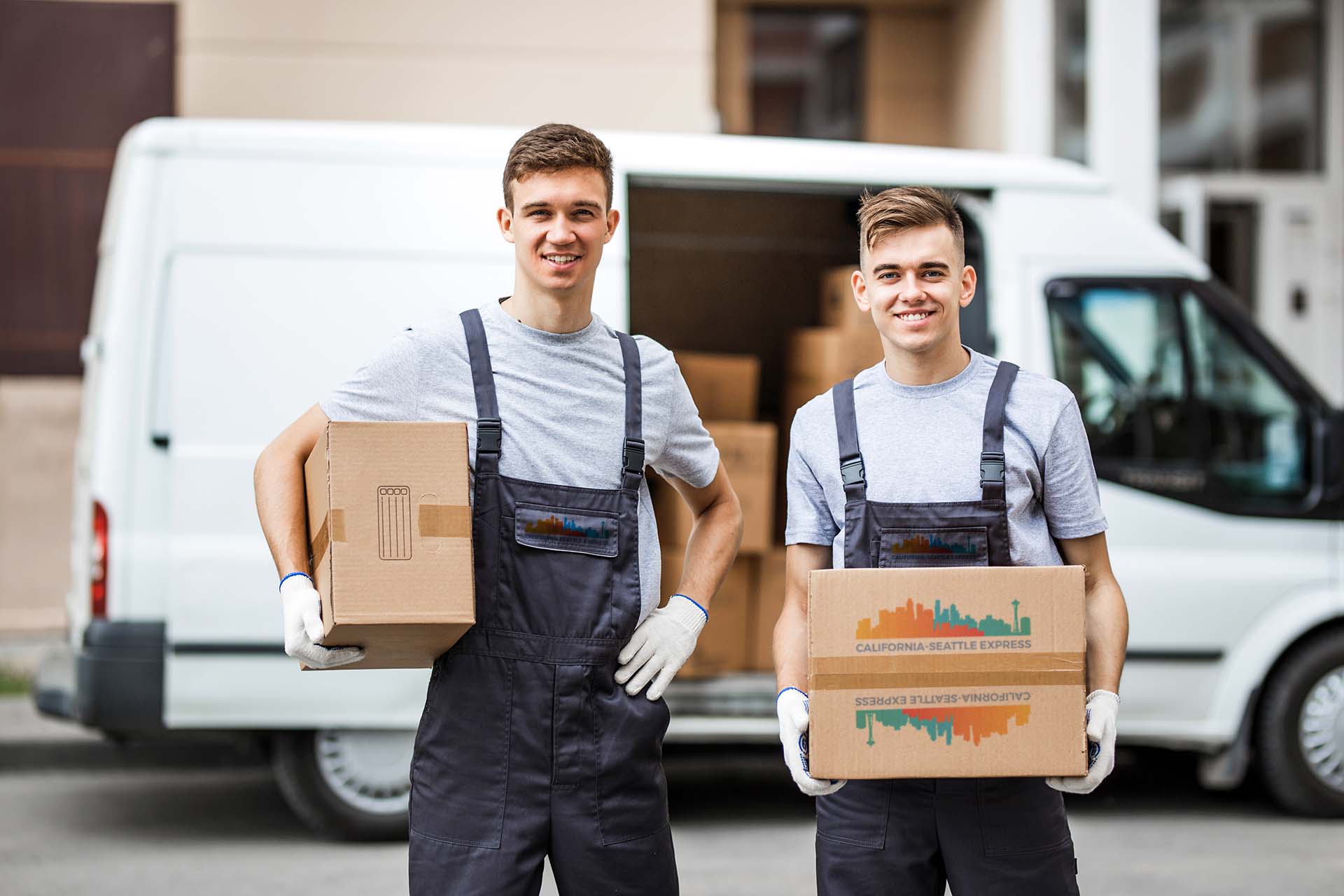 Two young handsome smiling workers wearing uniforms are standing next to the van full of boxes holding boxes in their hands. House move, mover service.