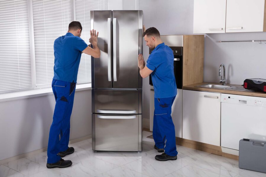 Two Young Male Movers Placing Steel Refrigerator In Kitchen